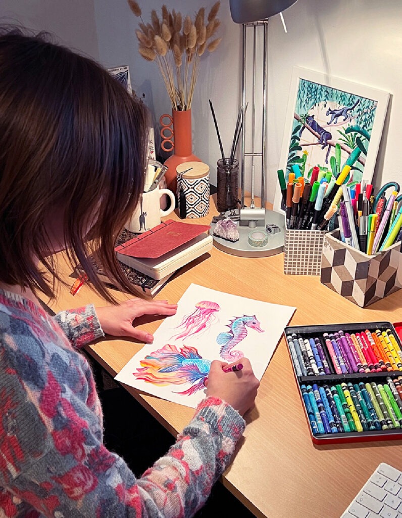 Photo de Sophie Bortier en pleine création, concentrée sur un dessin. Elle utilise des pastels à la cire sur une feuille de papier, entourée de son matériel artistique. L'image capture l'instant où l'art prend vie, reflétant son style poétique et sa passion pour le fait main.