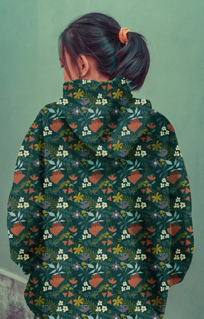 Femme vue de dos portant un sweat à capuche orné d’un motif fleuri vectoriel dans des tons verts foncés, rouges et jaunes. L’atmosphère est douce et moderne.